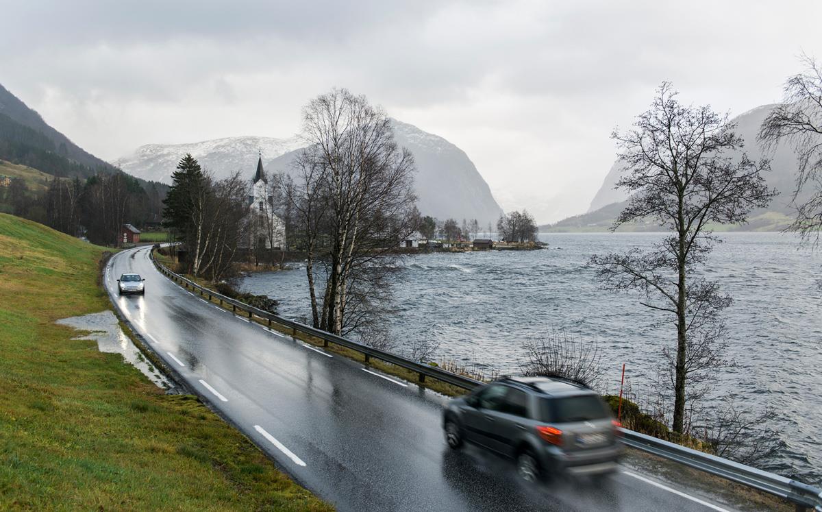 Foto av biler som kjører på våt vei langs et vann.