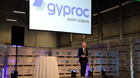 Statsministeren står på scenen hvor han holder åpningstalen. Gyproc-banner henger bak ham.