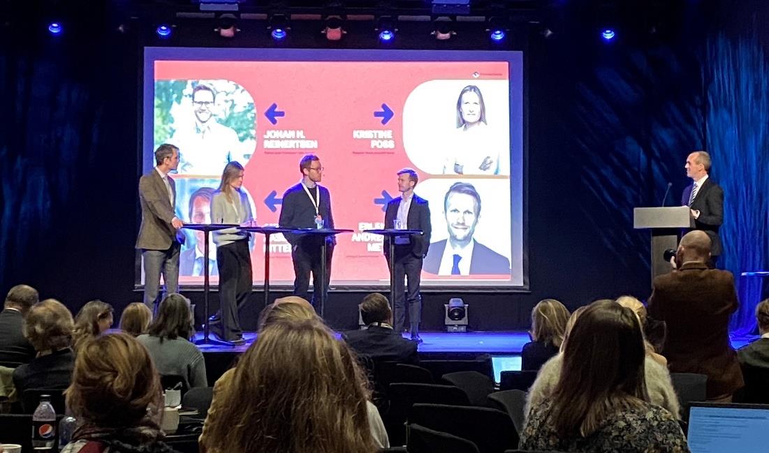 Fire paneldeltakere står bak små bord på et podium, sammen foran en powerpoint-presentasjon. Moderator står bak en talerstol til høyre, engasjert i samtale med paneldeltakerne. Foto: Kontaktpunktet