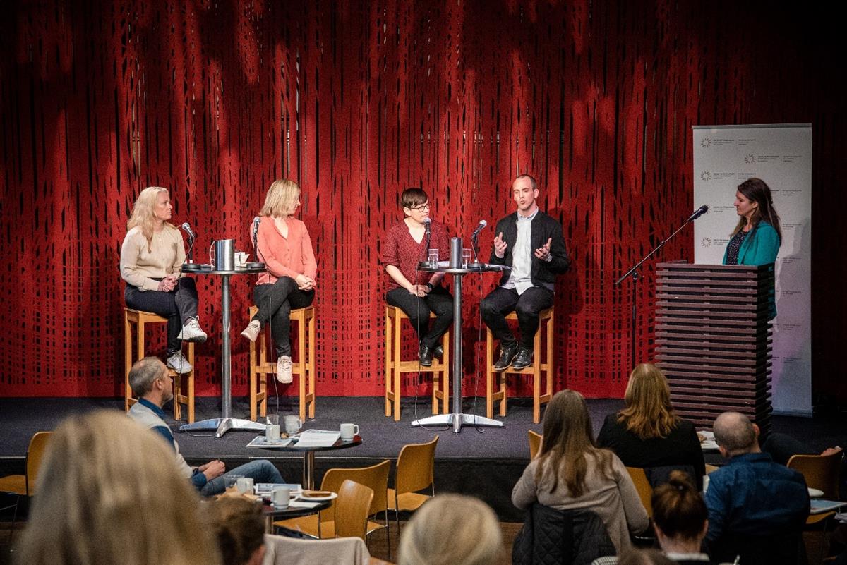 Fem mennesker sitter på et podium. En mann snakker og gestikulerer, mens de andre følger interessert med. Foto: Thorbjørn Helin