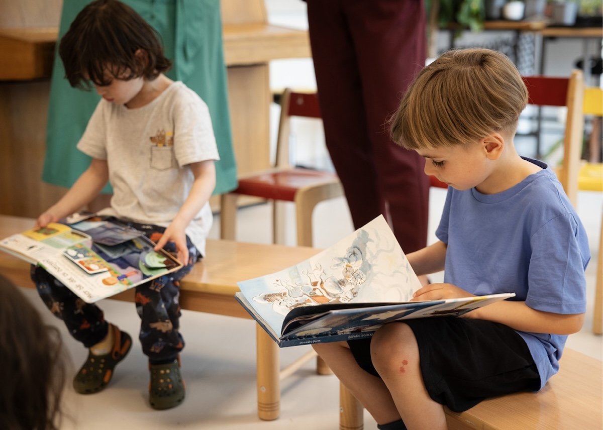 To barn sitter på hver sin benk i en barnehage. De leser hver sin bok