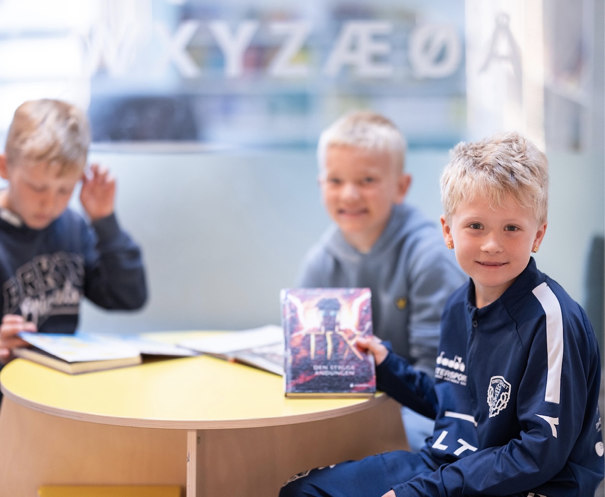 Tre barn sitter rundt et bord i et bibliotek