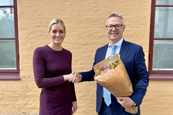 Kvinne i mørk kjole og mann i blå dress tar hverandre i hendene og smiler mot kamera. Mannen holder en bukett med blomster. De står foran en gul murvegg.