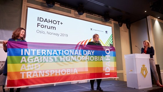 Minister of Culture and Equality Trine Skei Grande and UK Minister of State for Equalities Baroness Williams of Trafford on stage holding a big rainbow banner between them. 