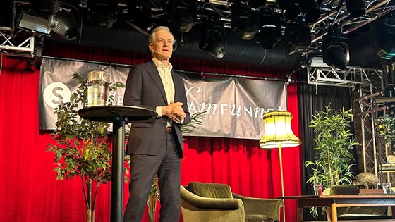 Jonas Gahr Støre står på en scene og holder tale. Rød gardin i bakgrunnen, og en Studentersamfunnet-logo på banner trukket over scenen.