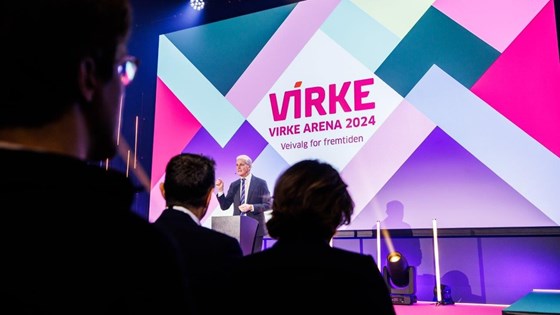 Statsministeren står bak en talerstol på en scene med stor Virke-logo på lerret i bakgrunnen.