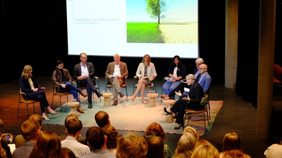 Bilde av et panel på ni personer sittende på stoler på scene